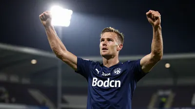 A tight action shot of a Bodø/Glimt player, mid-20s, athletic build, wearing a dark blue jersey, celebrating a goal with...