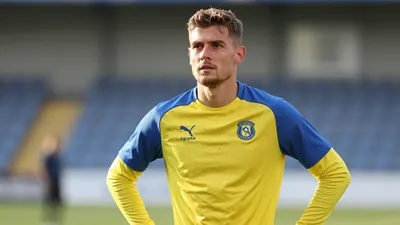 A midfield player in his late 20s, dressed in a plain yellow and blue Lillestrøm SK training top, stands pitchside at...