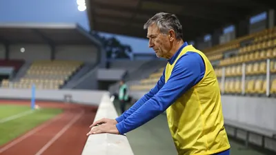 A 50-year-old male assistant coach with a weathered face, dressed in a plain yellow and blue polyester training top...