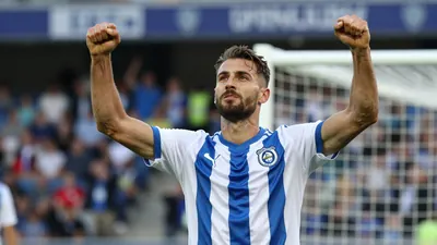 A Lillestrøm SK player in a blue and white jersey, mid-30s, muscular build, celebrating with arms raised after a goal in...
