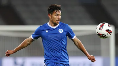 A mid-30s anonymous Lillestrøm SK midfielder in a solid blue kit, waist-up, focused on the ball as he prepares to strike...