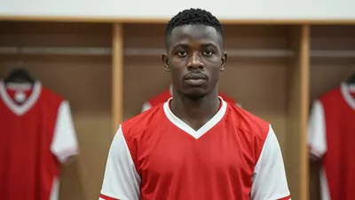 Photorealistic portrait of an 18-year-old Nigerian winger in a red and white football kit, standing in a locker room with...
